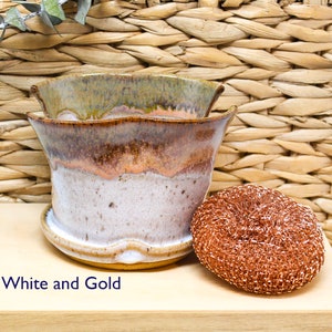 May include: White and gold ceramic planter with a wavy rim. The planter is sitting on a wooden surface with a copper scouring pad next to it.