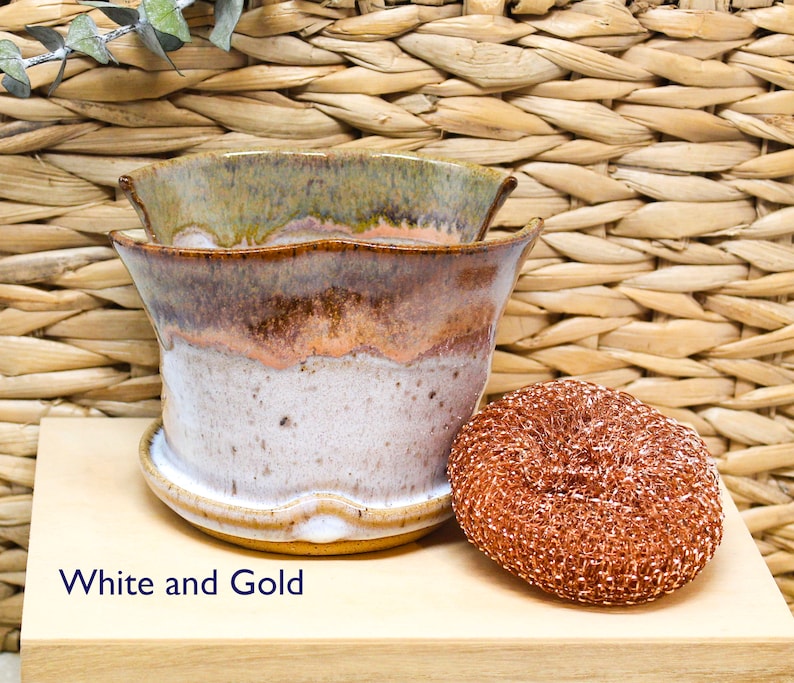 May include: White and gold ceramic planter with a wavy rim. The planter is sitting on a wooden surface with a copper scouring pad next to it.