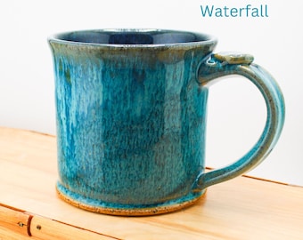 May include: A teal blue ceramic mug with a brown rim and handle. The mug has a speckled glaze and the word "Waterfall" is written in blue text on a white background.