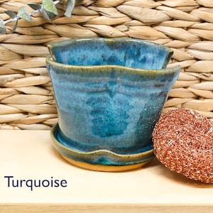 May include: Turquoise ceramic planter with a wavy rim and a light brown base. The planter is sitting on a wooden surface with the word "Turquoise" written in blue.