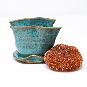 May include: A teal blue ceramic dish with a wavy rim and a matching saucer. The dish is next to a copper-colored scouring pad.