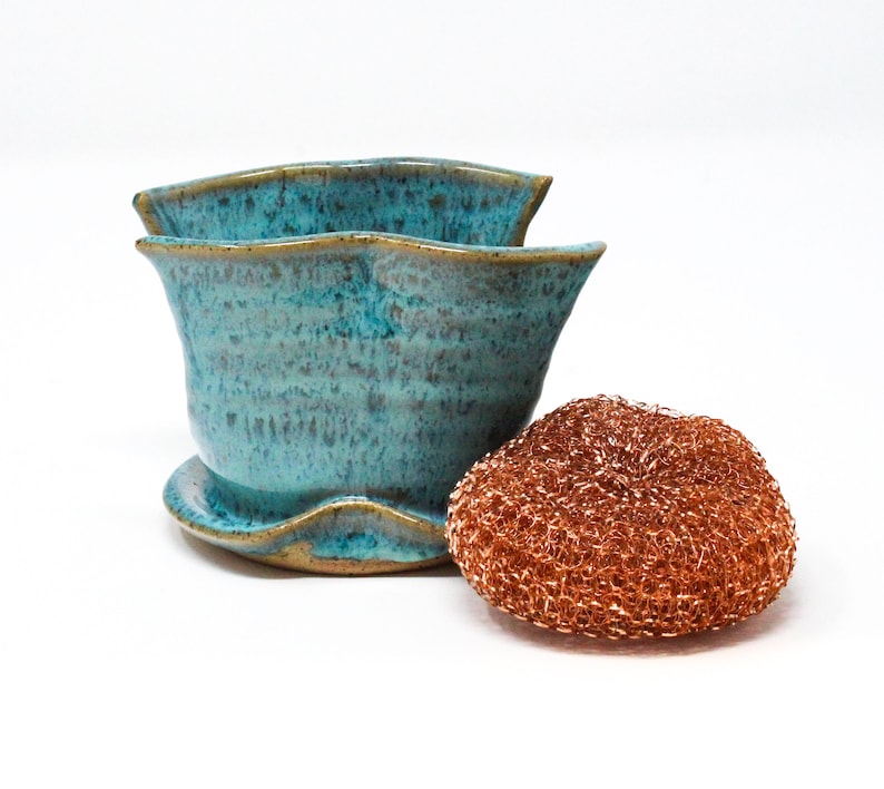 May include: A teal blue ceramic dish with a wavy rim and a matching saucer. The dish is next to a copper-colored scouring pad.