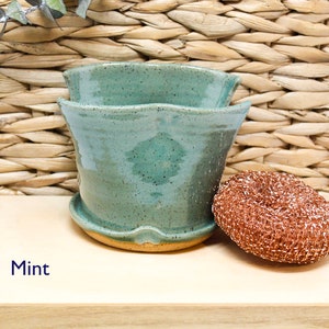 May include: A mint green ceramic planter with a wavy rim. The planter is sitting on a wooden surface with a copper-colored scouring pad next to it. The text "Mint" is written on the surface.