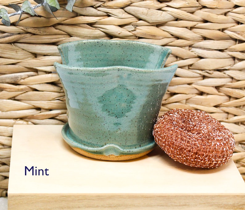 May include: A mint green ceramic planter with a wavy rim. The planter is sitting on a wooden surface with a copper-colored scouring pad next to it. The text "Mint" is written on the surface.