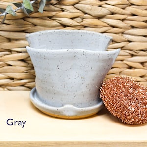 May include: Gray ceramic planter with a speckled glaze. The planter has a wide, rounded base and a flared lip. The planter is sitting on a wooden surface.
