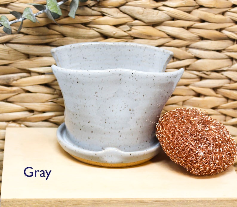 May include: Gray ceramic planter with a speckled glaze. The planter has a wide, rounded base and a flared lip. The planter is sitting on a wooden surface.