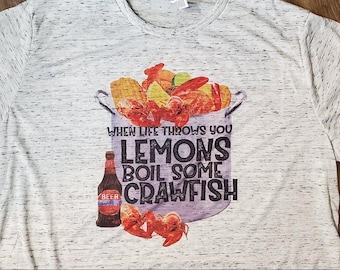 May include: A light gray t-shirt with a graphic of a pot filled with crawfish, corn, and lemons. The text reads "When life throws you lemons, boil some crawfish." A beer bottle is also featured.