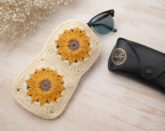 May include: A cream-colored crochet glasses case featuring two prominent yellow sunflower motifs with brown centers. The case is positioned next to a pair of black-framed sunglasses and a black leather Ray-Ban branded glasses case.