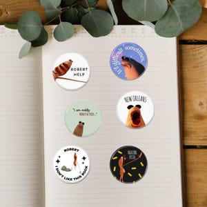 May include: A collection of six round pinback buttons with humorous illustrations and text. The buttons feature images of animals and food with phrases like "ROBERT HELP", "It's difficult sometimes", and "I don't like this rock".