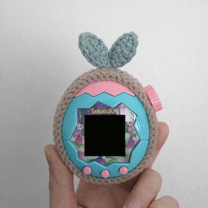 May include: A teal Tamagotchi virtual pet toy with a pink button and a black screen, encased in a crocheted brown cover with blue bunny ear accents. The text "Tamagotchi" is visible on the device.