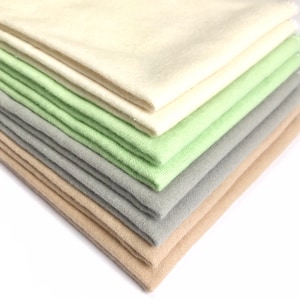 Op de afbeelding: Een stapel stofstalen in verschillende tinten beige, groen en grijs. De stof is zacht en heeft een textuur, en de kleuren zijn gedempt en kalmerend.