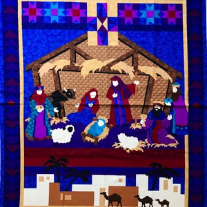 Pode incluir: Um painel de tecido azul com um design acolchoado que representa a cena da natividade. A cena inclui um estábulo, uma manjedoura, um bebê Jesus, Maria, José e os três reis magos. O fundo é um céu azul com estrelas. A parte inferior do painel apresenta uma paisagem marrom e branca com palmeiras e camelos.