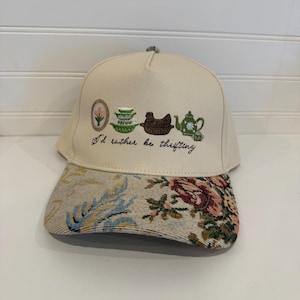Peut inclure: Une casquette de baseball couleur crème avec une broderie représentant une fleur encadrée, des bols verts empilés, un plat en forme de poule marron et une théière verte. Sous la broderie, le texte indique I'd rather be thrifting. La visière a un motif floral.