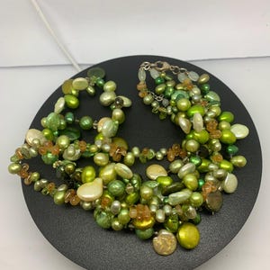 Peut inclure: Un collier à plusieurs rangs composé de perles et de perles de couleur verte et blanc cassé. Le collier comprend un mélange de formes et de tailles, avec quelques accents de couleur ambre. Le collier est présenté sur une surface noire.