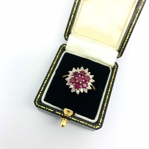 May include: A gold ring with a cluster of red rubies surrounded by diamonds. The ring is in a black velvet ring box.