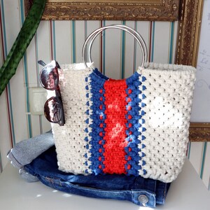 May include: A white crocheted tote bag with blue and red stripes. The bag has a silver ring handle and is sitting on top of a pair of blue jeans.