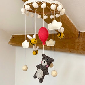 May include: A white felt baby mobile with a wooden ring, white pom-poms, and felt clouds, bees, a red balloon, and a gray bear.