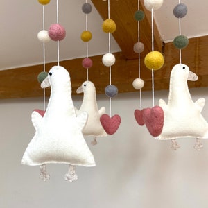 Pode incluir: Um móbile de feltro branco com três gansos, corações cor-de-rosa e bolas de feltro coloridas. Os gansos têm olhos verdes e detalhes cor-de-rosa. O móbile está pendurado em uma viga de madeira.