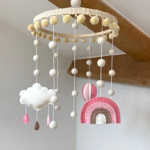 May include: A white felt baby mobile with a pink and brown rainbow, a white cloud with pink and brown raindrops, and a hot air balloon. The mobile is hanging from a wooden hoop with white pom-poms around the edge.