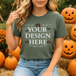 May include: A green t-shirt with the text "YOUR DESIGN HERE" in white, decorated with bats and pumpkins. The shirt is paired with blue jeans. The background features carved pumpkins and hay bales, creating a festive autumn scene.