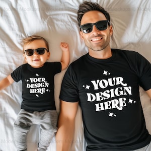 May include: A black t-shirt with the text "YOUR DESIGN HERE" in white, worn by an adult and a child, both wearing sunglasses. The child is also wearing gray sweatpants. The image is taken from above.