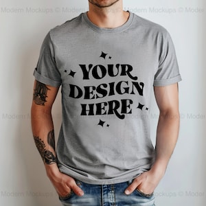 Könnte beinhalten: Ein graues T-Shirt mit einer schwarzen Grafik, auf der in einer Retro-Schrift "YOUR DESIGN HERE" steht. Die Grafik ist von kleinen Sternen umgeben.