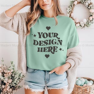 May include: A woman wearing a light green t-shirt with the text "YOUR DESIGN HERE" in black. The shirt has a crew neck and short sleeves. The woman is wearing a light brown cardigan and blue denim shorts.