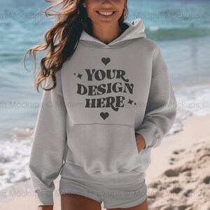 May include: A gray hooded sweatshirt with the text "YOUR DESIGN HERE" in black lettering and a small heart graphic. The sweatshirt is being worn by a person standing on a beach.