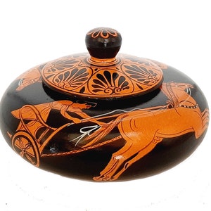 Puede incluir: Una caja de madera negra con una tapa decorada con diseños griegos de color naranja y negro. La tapa presenta una escena de carroza estilizada con caballos y un conductor. La caja es un contenedor de almacenamiento decorativo.
