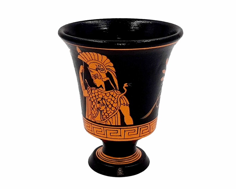 Pu&ograve; includere: Bicchiere da bere in ceramica nera e arancione con un design greco che raffigura un guerriero con elmo e scudo. Il bicchiere ha una base larga e una parte superiore stretta.