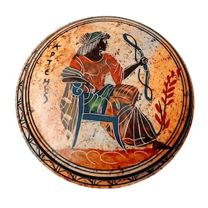 Puede incluir: Un plato redondo de cerámica con una imagen pintada de una mujer sentada en una silla. La mujer lleva una túnica roja y azul y sostiene un bastón. El plato tiene un fondo marrón con un borde negro y la palabra "Artemisa" escrita en letras griegas.