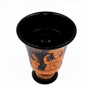 Pu&ograve; includere: Una tazza in ceramica nera e arancione con un design greco. La tazza presenta una donna con un elmo e un uccello su una colonna.