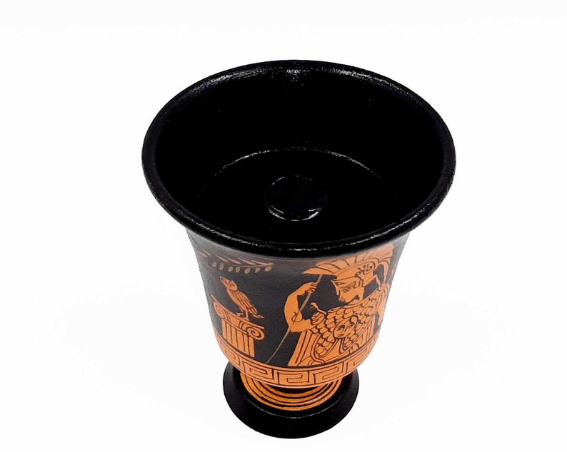Pu&ograve; includere: Una tazza in ceramica nera e arancione con un design greco. La tazza presenta una donna con un elmo e un uccello su una colonna.