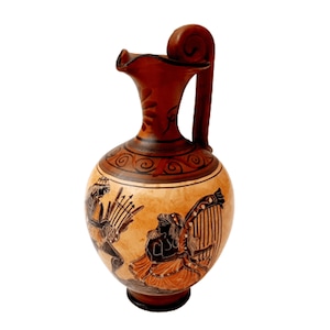 Puede incluir: Una jarra de cerámica marrón con un diseño pintado en blanco y negro de dos figuras tocando instrumentos musicales. La jarra tiene un asa curva y un cuerpo redondo.