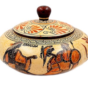 May include: A round ceramic trinket box with a lid, decorated with a black and orange Greek-style design. The box features a scene of two figures with spears and shields, and the word "APOLLON" is written on the side. The lid is decorated with a floral pattern.