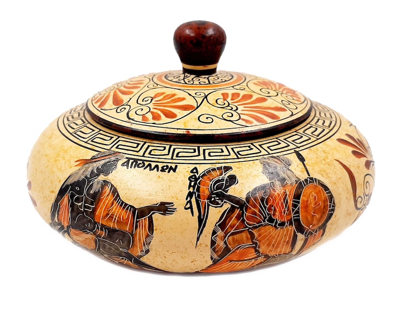 May include: A round ceramic trinket box with a lid, decorated with a black and orange Greek-style design. The box features a scene of two figures with spears and shields, and the word "APOLLON" is written on the side. The lid is decorated with a floral pattern.