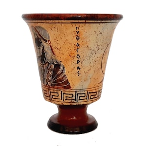 May include: A terracotta drinking cup with a black and white design of a woman's head and Greek writing. The cup has a round base and a flared top.