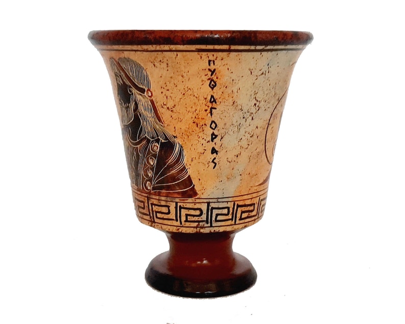 May include: A terracotta drinking cup with a black and white design of a woman's head and Greek writing. The cup has a round base and a flared top.