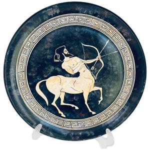 May include: A circular, dark blue decorative plate with a white centaur figure aiming a bow and arrow. The plate has a white geometric border design and is displayed on a white stand. The artwork is reminiscent of ancient Greek pottery.