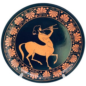 May include: A circular, black decorative plate with an orange centaur playing a wind instrument. The plate has an orange floral border design. The centaur is the central focus of the plate, set against a white background.