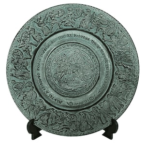 May include: A round, green metal plate with a raised design depicting a scene from Greek mythology. The plate has a Greek inscription around the edge and is on a black stand.