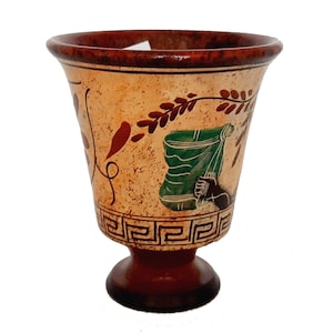 May include: A small, brown ceramic cup with a white base and a decorative design of green, red, and brown leaves and a geometric pattern around the top.