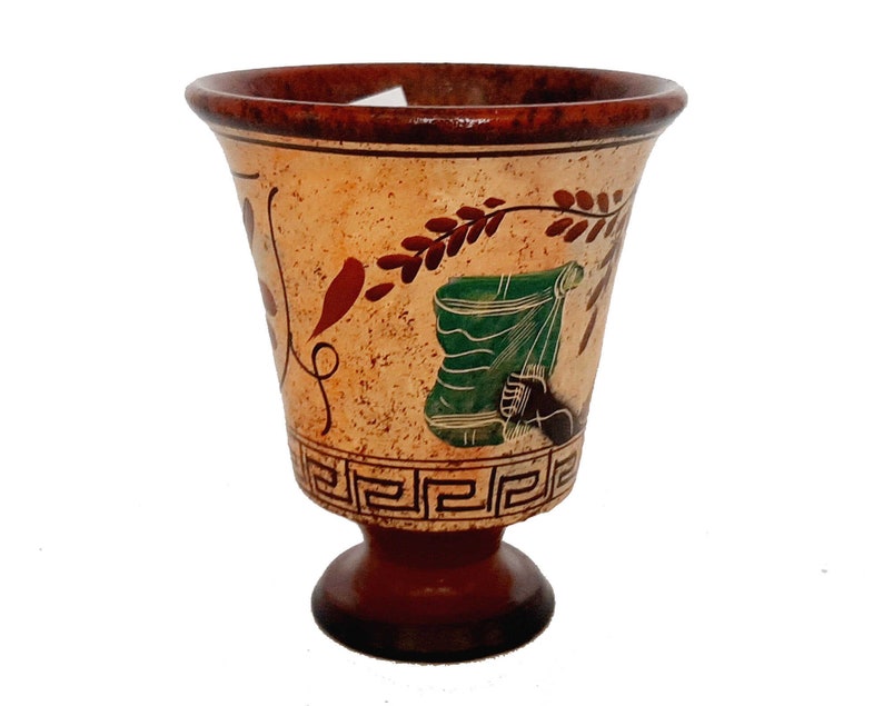 May include: A small, brown ceramic cup with a white base and a decorative design of green, red, and brown leaves and a geometric pattern around the top.