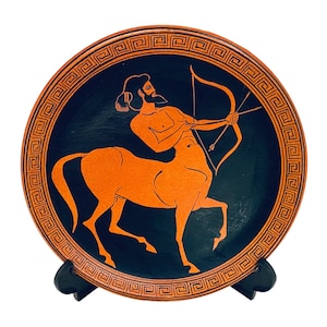 May include: A ceramic plate with a black background and a red-orange image of a centaur holding a bow and arrow. The plate has a decorative border of a repeating Greek key pattern.
