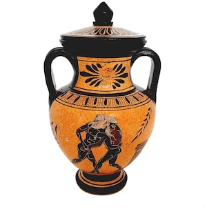 May include: A black and orange ceramic amphora with a lid, featuring a black and white painted design of two figures wrestling. The amphora has two handles and a round base.