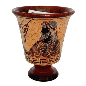 May include: A small, brown ceramic cup with a black and white design of a man's head and a vine. The cup has a base with a Greek key pattern.