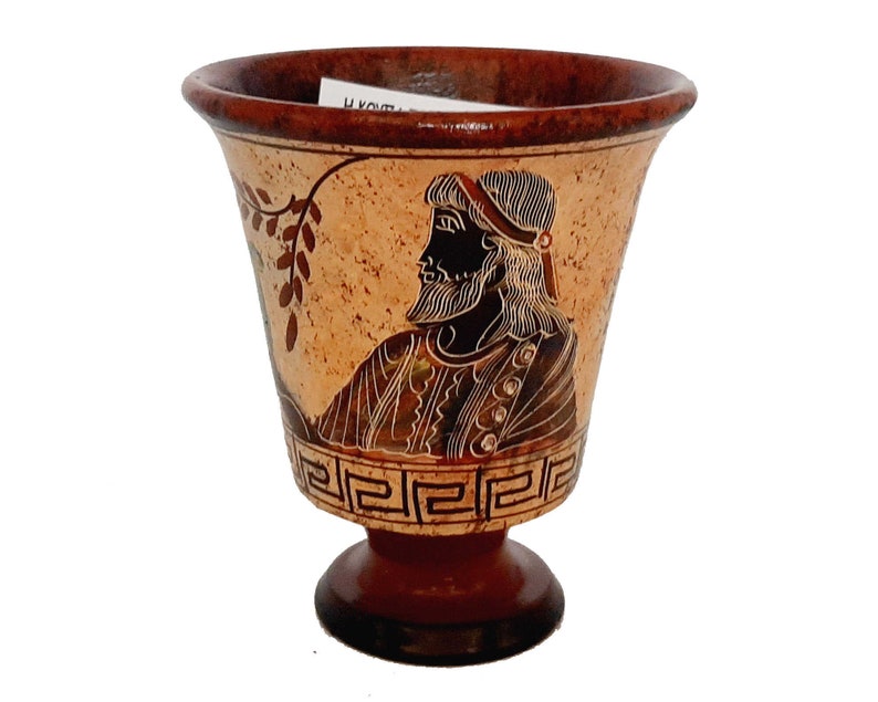 May include: A small, brown ceramic cup with a black and white design of a man's head and a vine. The cup has a base with a Greek key pattern.