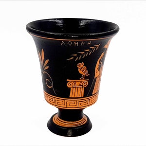 Pu&ograve; includere: Coppa da bere in ceramica nera con un design arancione. Il design presenta un gufo appollaiato su una colonna, un ramo con foglie e la parola "ATHENA" in lettere greche.