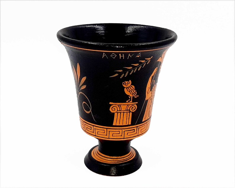 Pu&ograve; includere: Coppa da bere in ceramica nera con un design arancione. Il design presenta un gufo appollaiato su una colonna, un ramo con foglie e la parola "ATHENA" in lettere greche.