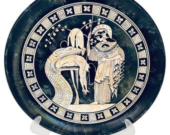 Pode incluir: Um prato azul escuro com uma borda branca e preta de chave grega e uma cena central. A cena retrata figuras e uma serpente em estilo clássico. O prato é exibido em um suporte transparente.